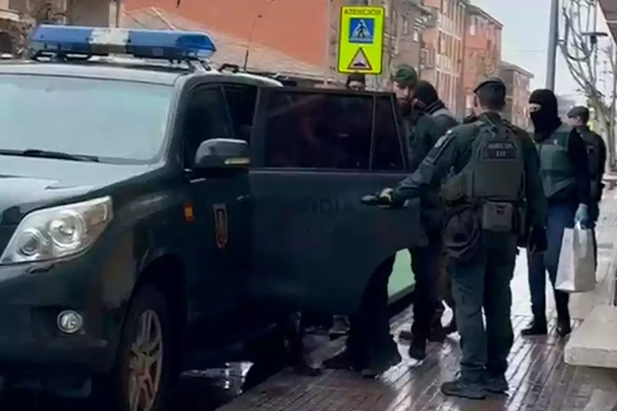 Un grupo de agentes de policía, algunos con pasamontañas, están junto a un vehículo policial en una calle urbana, mientras uno de ellos abre la puerta trasera del coche.