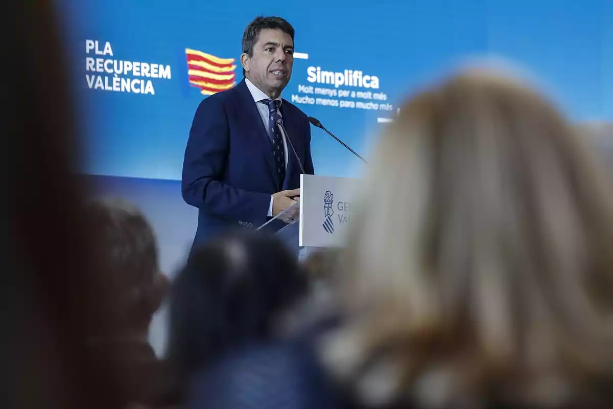 A man in a suit speaks at a podium during an event with a blue background displaying the text "PLA RECUPEREM VALÈNCIA" and "Simplifica."