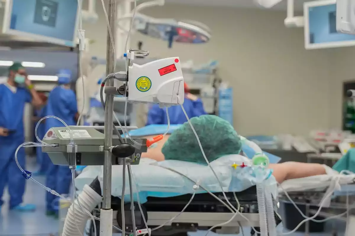 Paciente acostado en una camilla de hospital conectado a varios equipos médicos en un quirófano con personal de salud al fondo