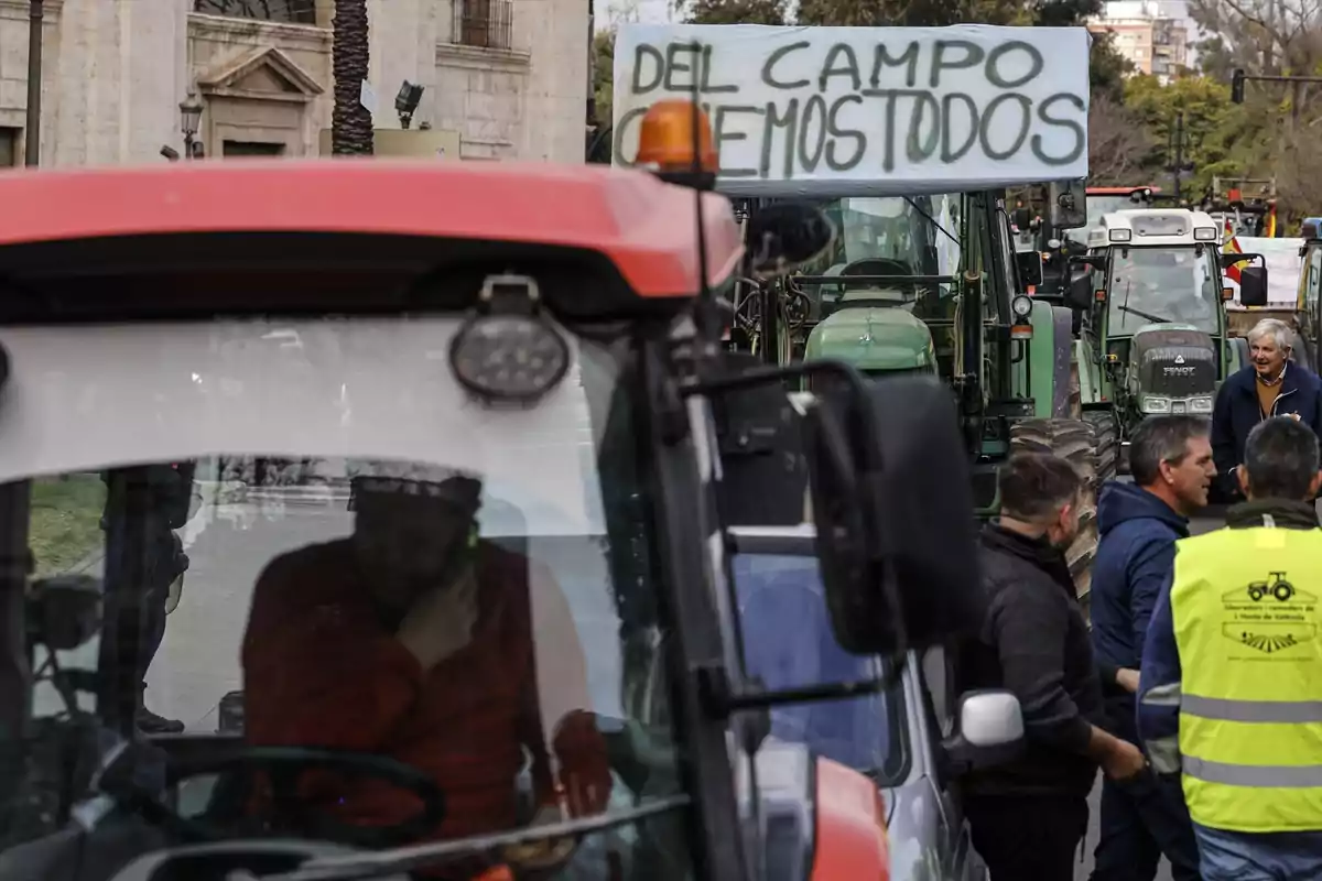 Un grupo de personas y tractores se encuentran en una calle durante una manifestación, con un cartel que dice "Del campo comemos todos".