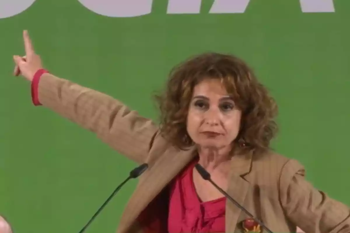 A woman in a beige jacket and red blouse gestures with her arm raised against a green background.