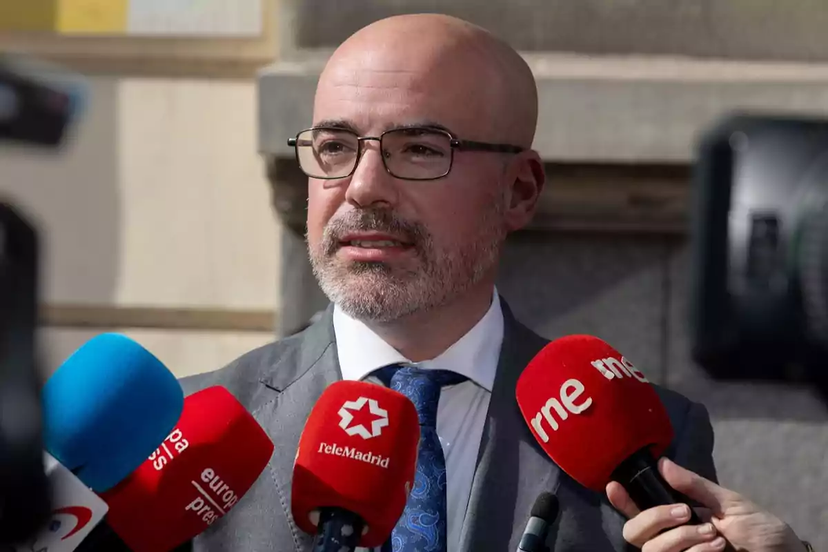 Un hombre con gafas y barba está hablando frente a varios micrófonos de diferentes medios de comunicación. Un hombre con gafas y barba está hablando frente a varios micrófonos de diferentes medios de comunicación.