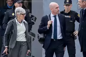 A group of people, including a woman with gray hair and a man in a suit, walk alongside police officers.