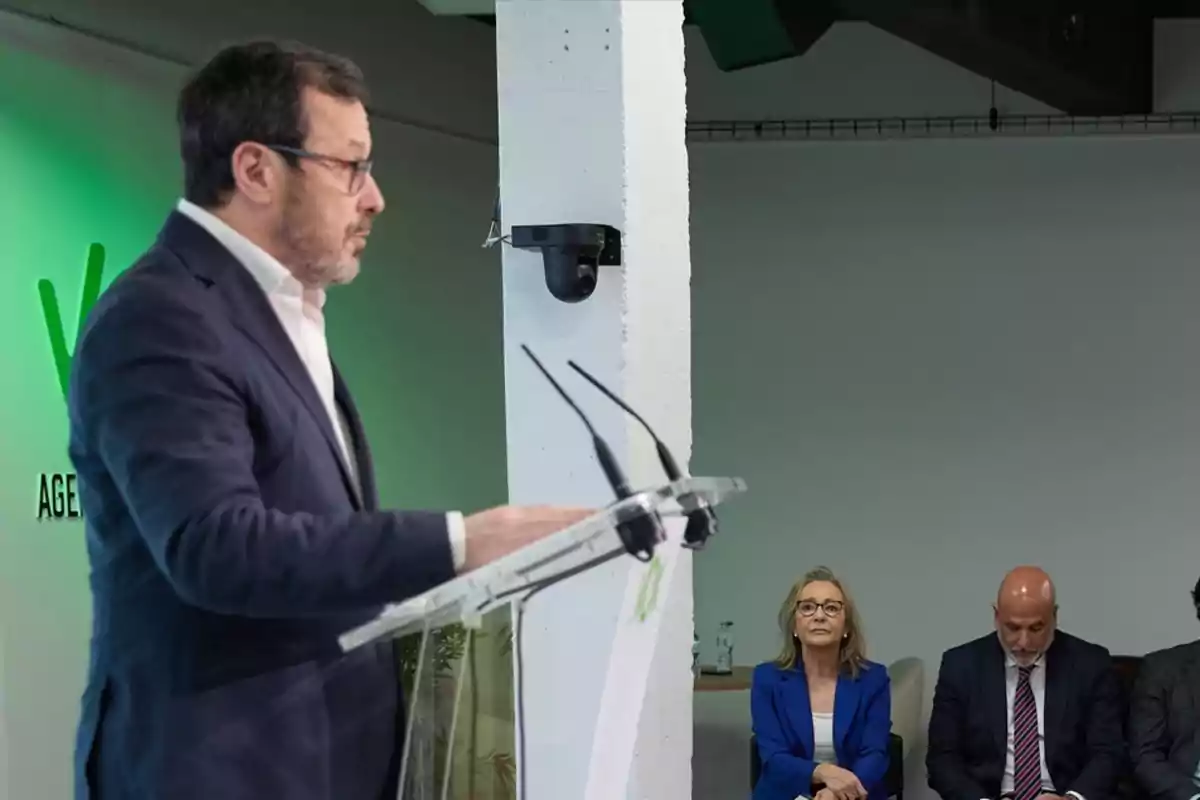Hombre de traje hablando en un atril mientras dos personas sentadas lo escuchan atentamente en un evento profesional Hombre de traje hablando en un atril mientras dos personas sentadas lo escuchan atentamente en un evento profesional