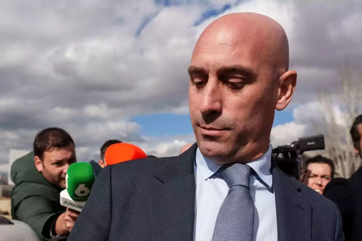 A bald man in a suit and tie is surrounded by colorful microphones while being interviewed outdoors under a cloudy sky.