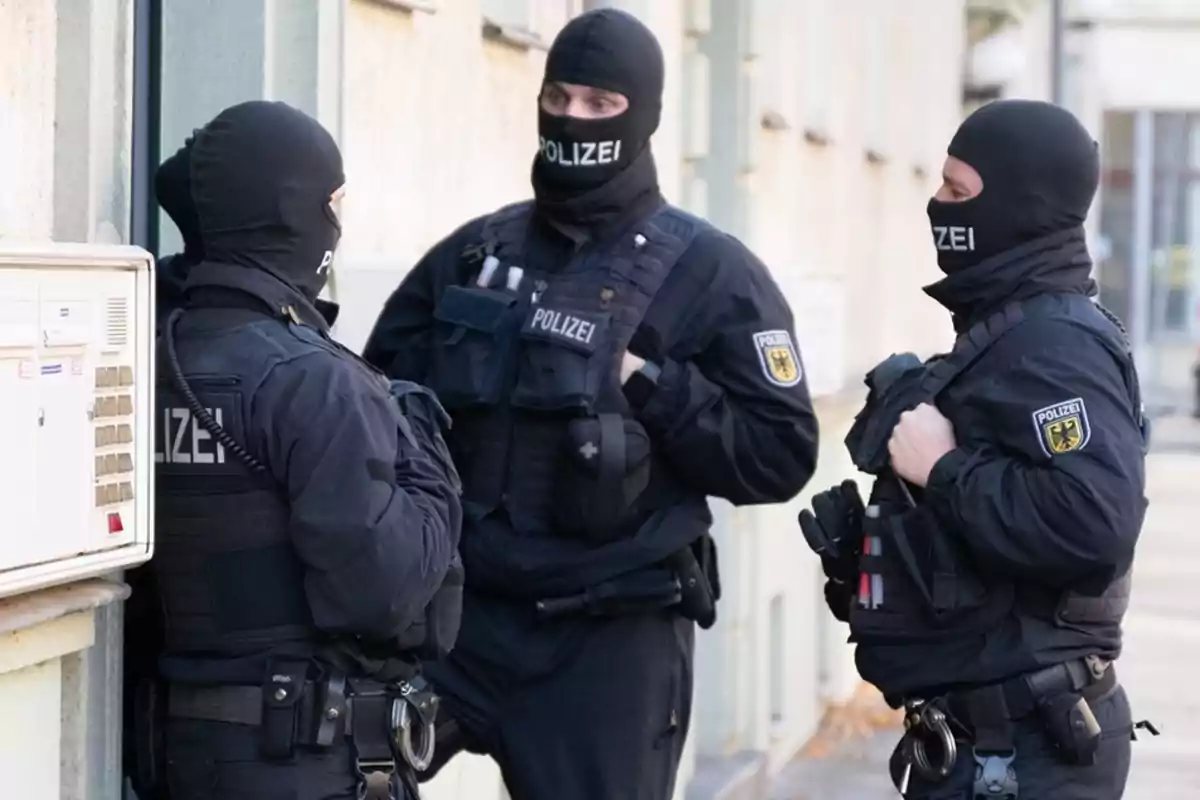 Tres agentes de policía con uniformes tácticos y pasamontañas conversan junto a un edificio.