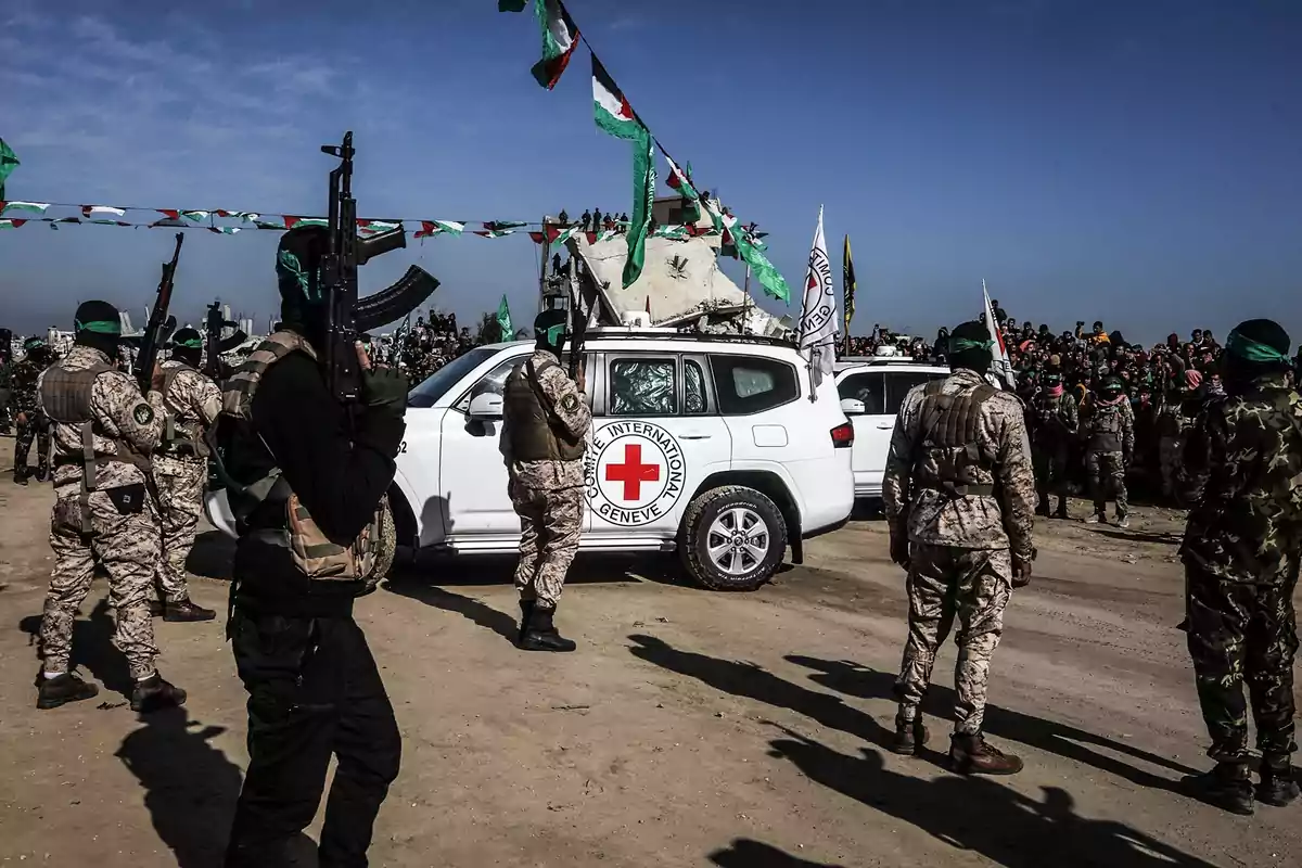 A group of armed individuals surrounds a white vehicle with the Red Cross emblem in an open area, while a crowd watches in the background.