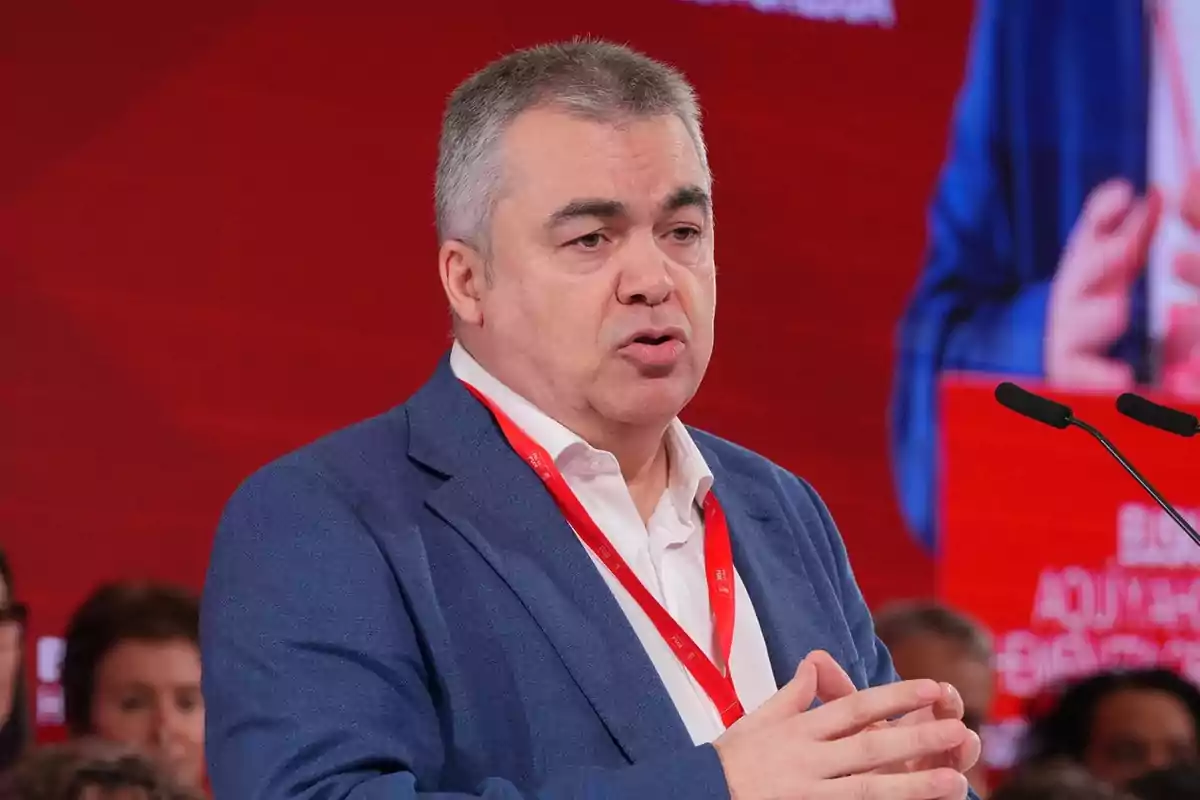Hombre de cabello canoso y saco azul hablando en un evento con fondo rojo y micrófonos frente a él Hombre de cabello canoso y saco azul hablando en un evento con fondo rojo y micrófonos frente a él