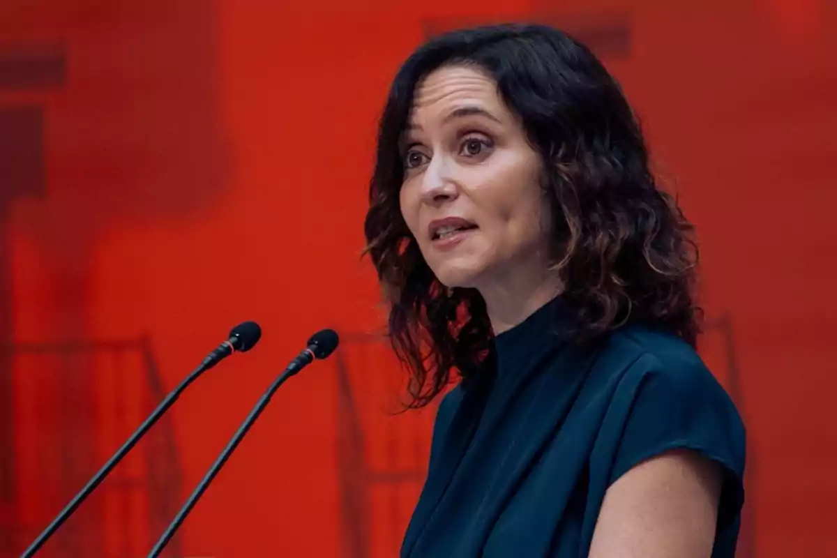 A woman speaking in front of two microphones with a red background.