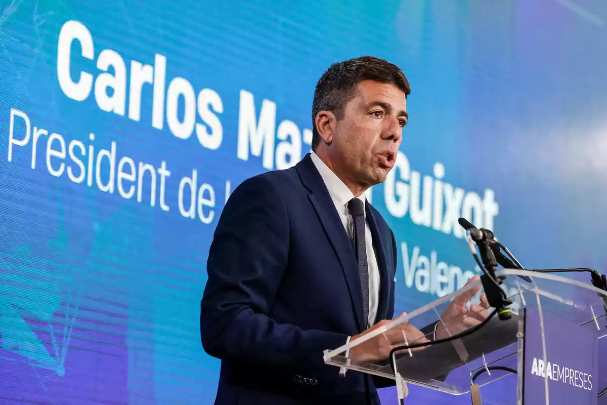 A man in a suit speaks at a podium with a microphone, in front of a blue screen with text.