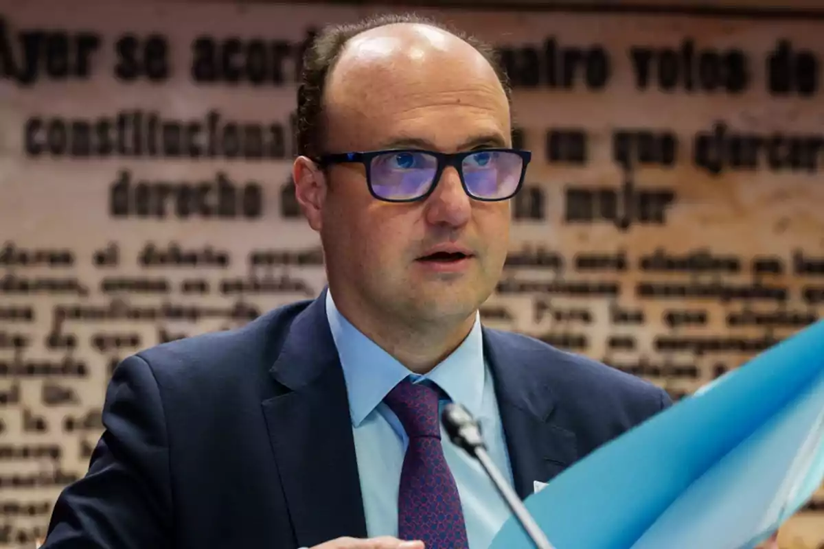 Un hombre con gafas y traje oscuro sostiene un documento azul mientras habla en un entorno formal con un fondo de texto borroso.