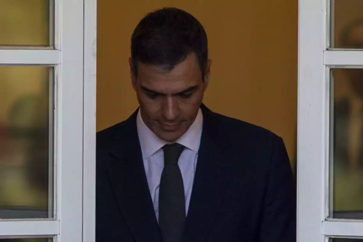 A man in a dark suit and tie looks down while standing in a doorway with a white frame.