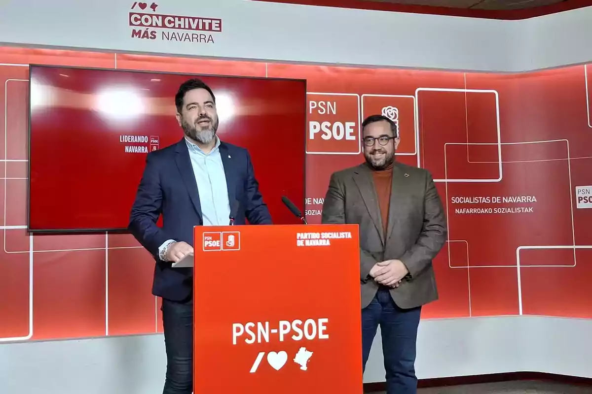 Dos hombres de pie en una sala de prensa del Partido Socialista de Navarra con fondo y atril rojos decorados con logotipos y mensajes del PSN-PSOE Dos hombres de pie en una sala de prensa del Partido Socialista de Navarra con fondo y atril rojos decorados con logotipos y mensajes del PSN-PSOE