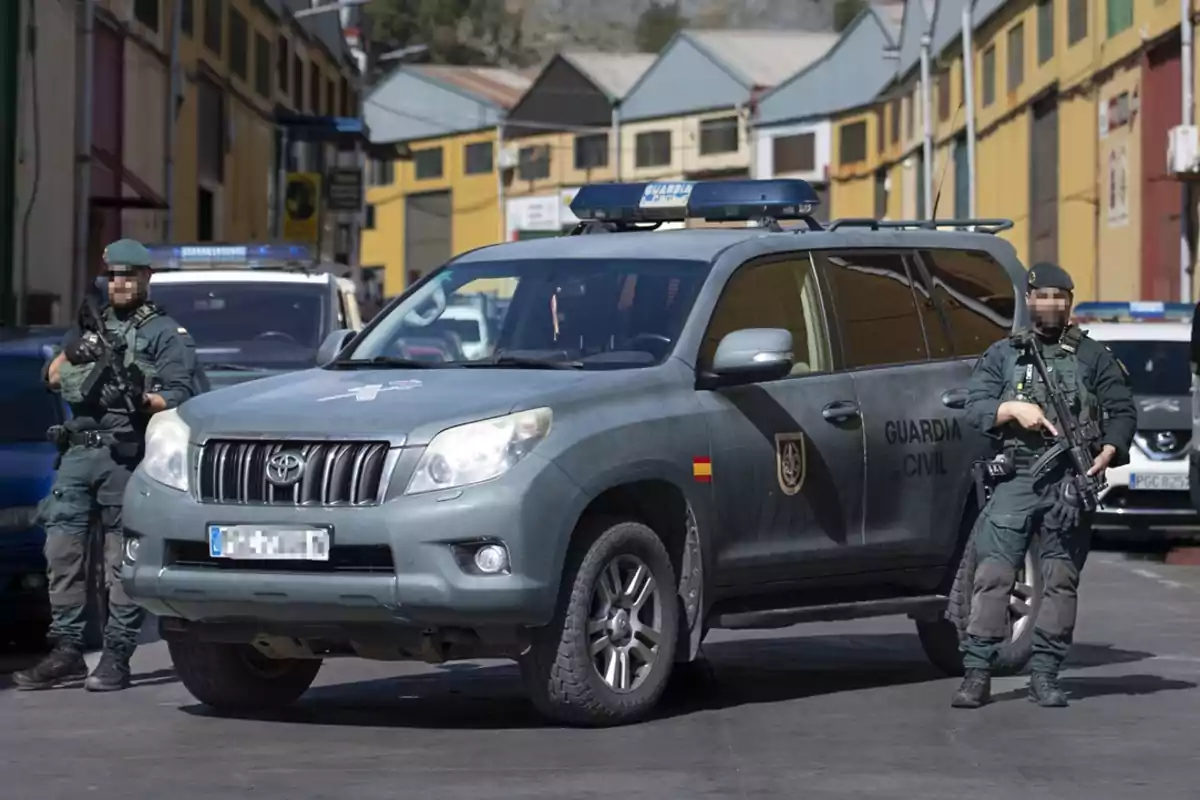 Un vehículo de la Guardia Civil con dos agentes armados de pie a su lado en una calle con edificios al fondo.