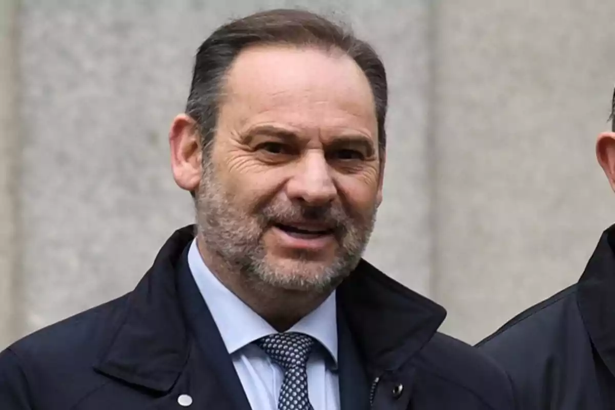 Hombre de mediana edad con barba y cabello corto vestido con chaqueta oscura y corbata sonríe frente a un fondo gris Hombre de mediana edad con barba y cabello corto vestido con chaqueta oscura y corbata sonríe frente a un fondo gris