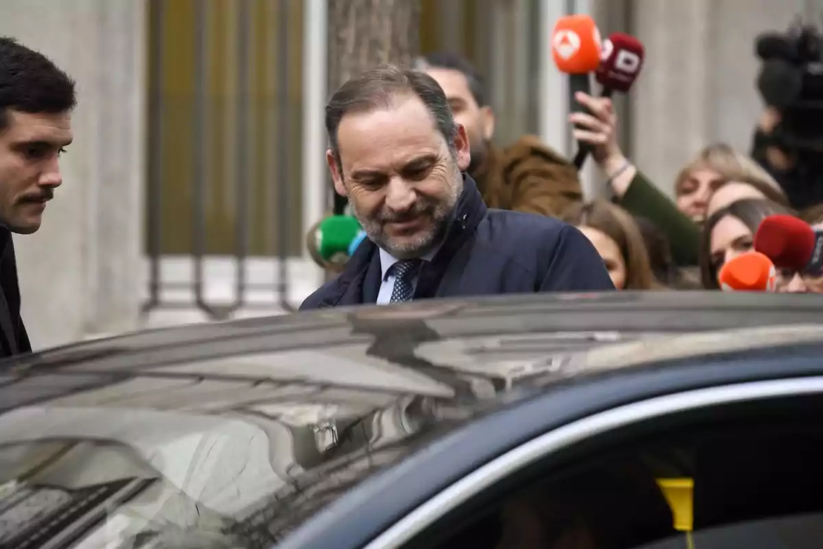 A man with a beard and a dark suit is surrounded by journalists with colorful microphones as he approaches a car.