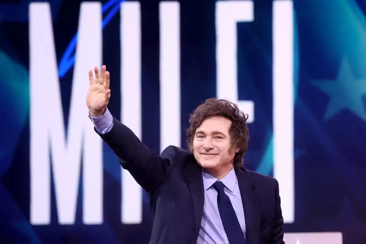 A man in a suit and tie raises his hand in greeting in front of a background with large letters.