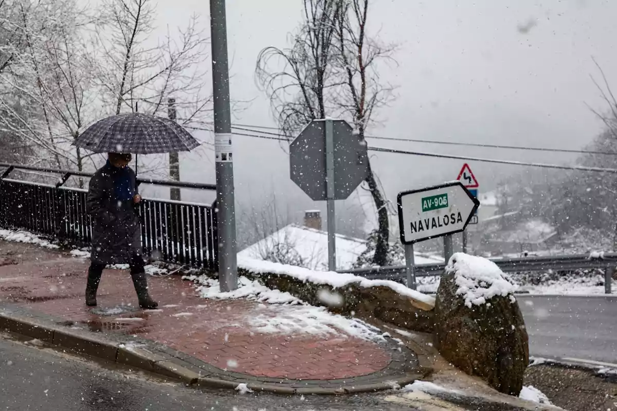 A person walks under the snow with an umbrella on a path next to a sign indicating the direction to Navalosa.