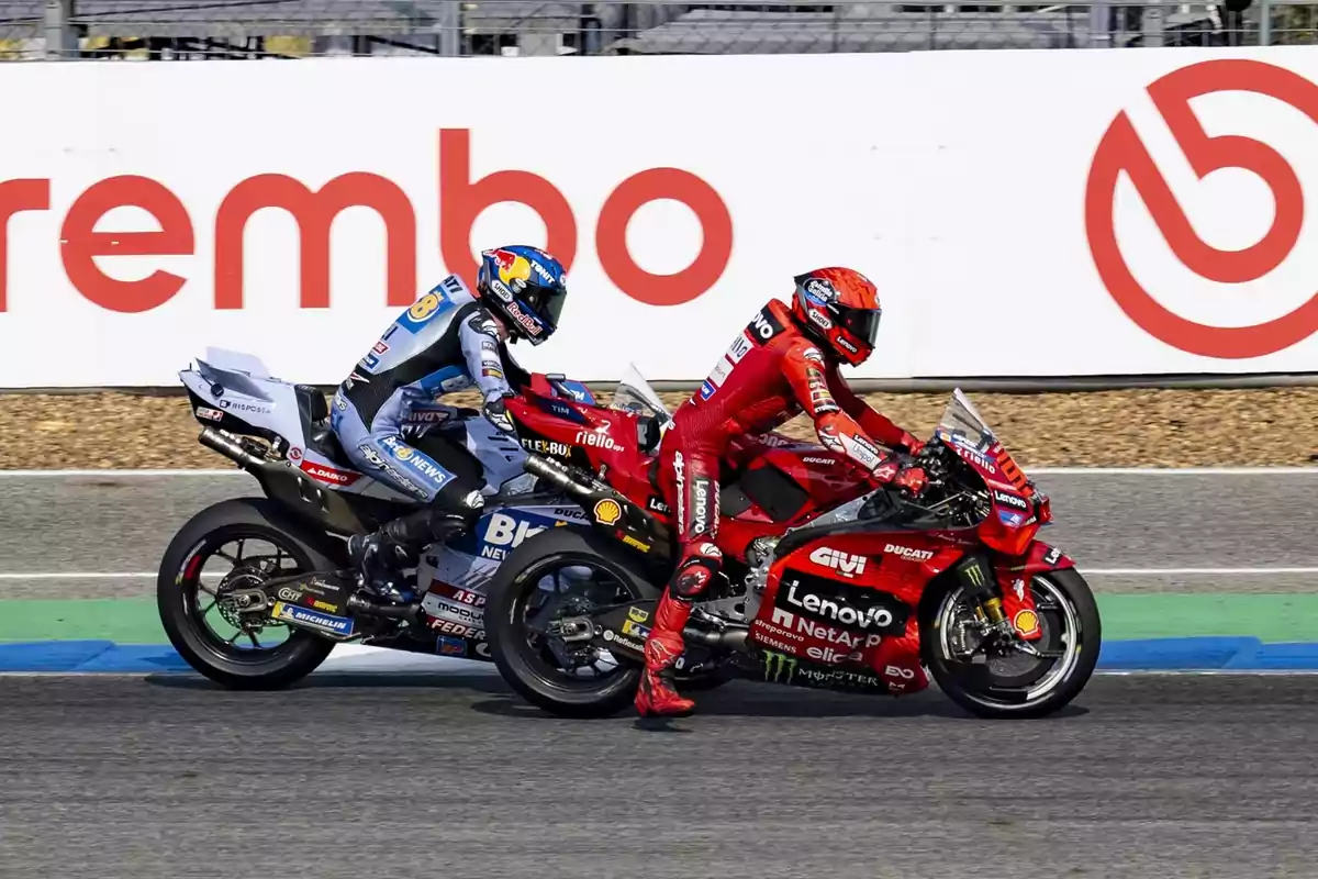 Dos motociclistas compiten en una carrera, uno con traje azul y otro con traje rojo, en un circuito con publicidad de Brembo al fondo.