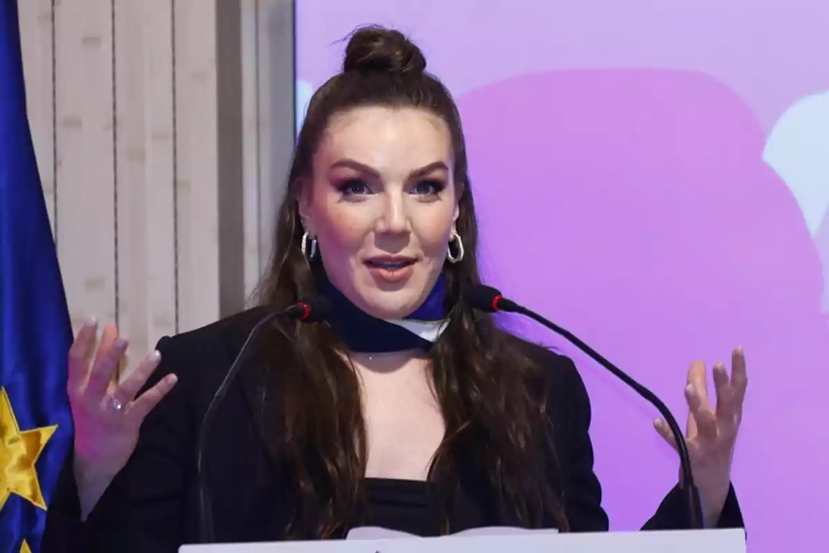 A person speaking at a podium with microphones and a purple background.