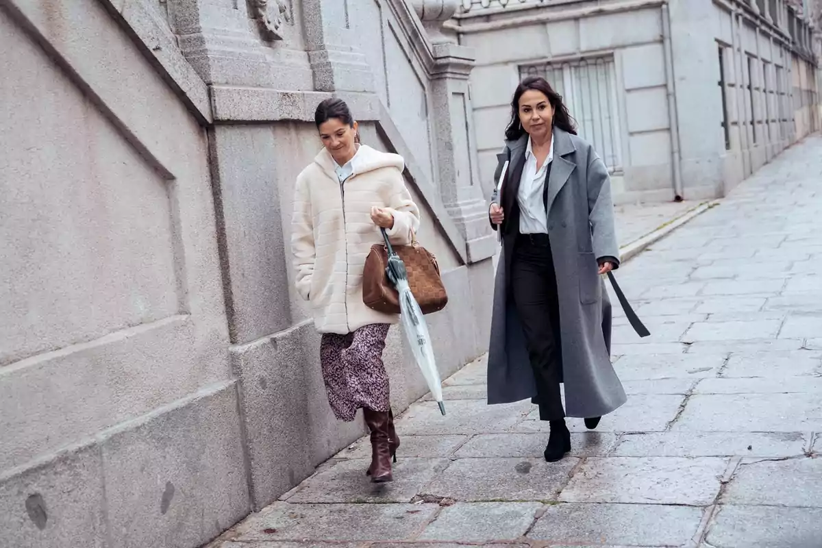 Dos mujeres caminan por una acera de piedra junto a un edificio de paredes grises, ambas llevan abrigos largos y una de ellas sostiene un paraguas transparente Dos mujeres caminan por una acera de piedra junto a un edificio de paredes grises, ambas llevan abrigos largos y una de ellas sostiene un paraguas transparente