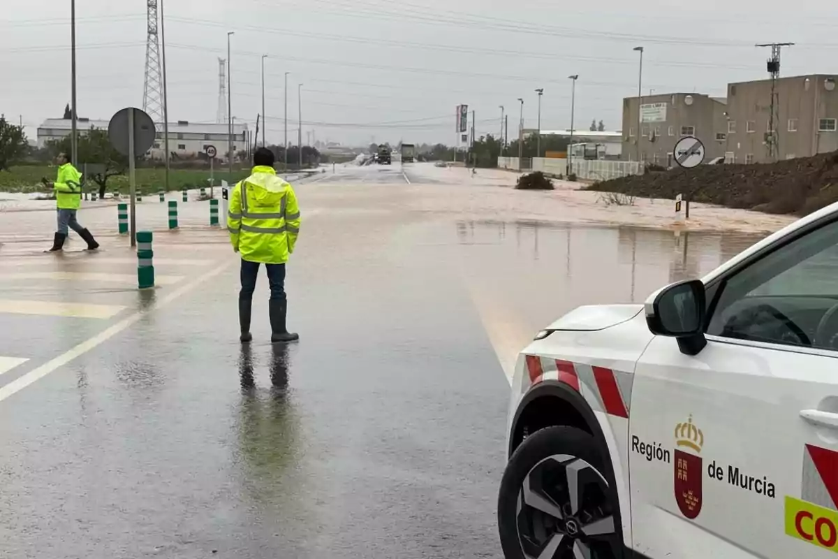 Personas con chaquetas reflectantes y botas de lluvia observan una carretera inundada en una zona industrial, mientras un vehículo oficial de la Región de Murcia está estacionado cerca.