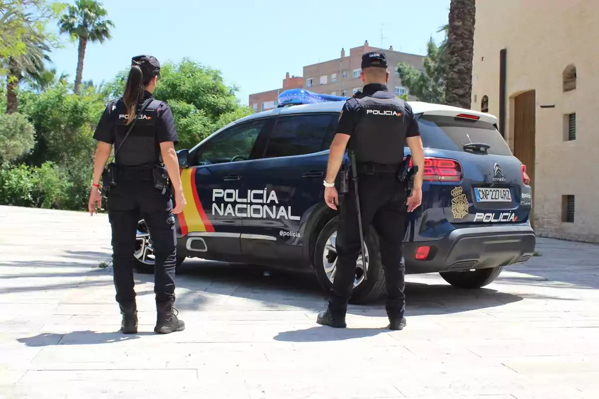 Dos agentes de la policía nacional de pie junto a un coche patrulla en un entorno urbano con vegetación y edificios al fondo.