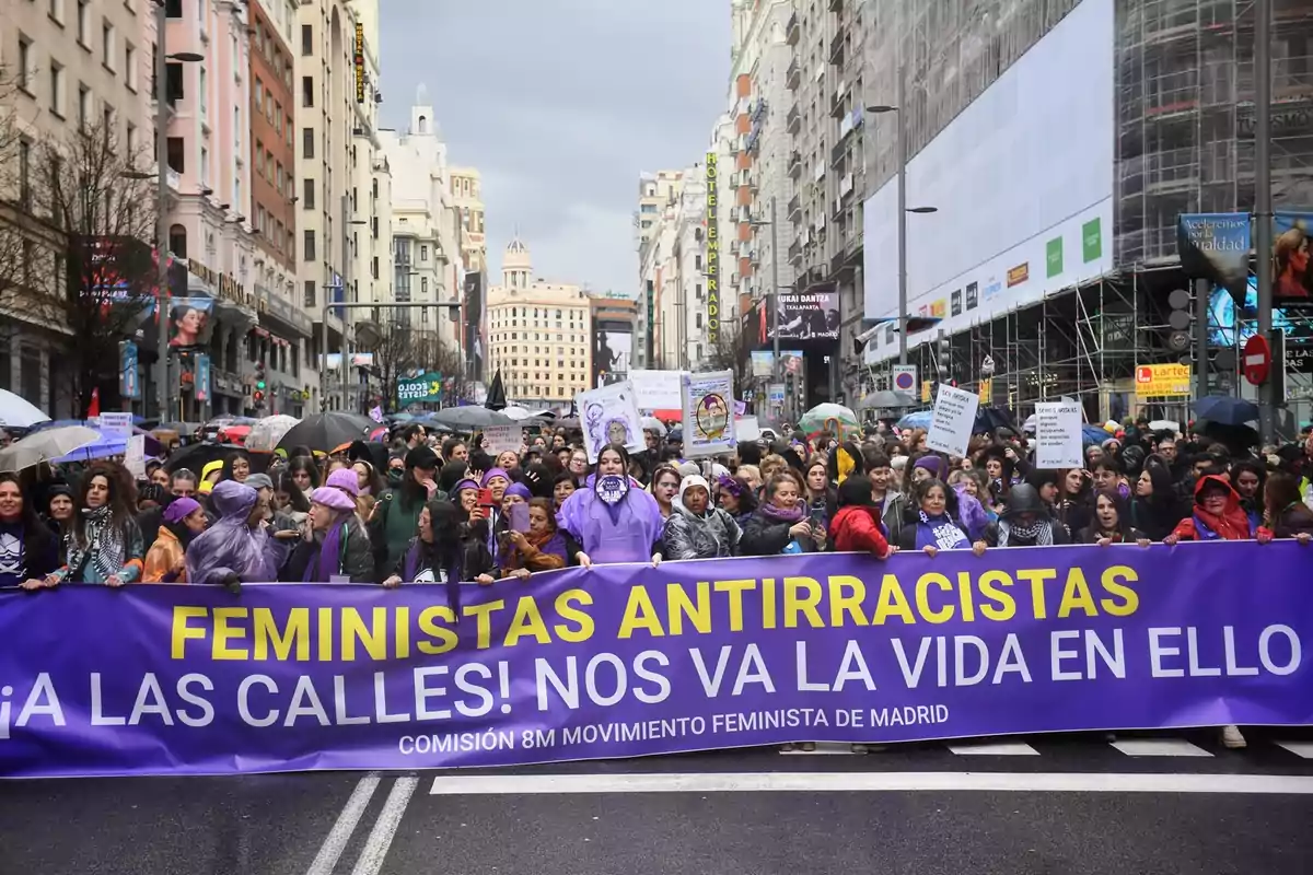 A crowd participates in a feminist demonstration on a street in Madrid, holding a banner that says "Anti-Racist Feminists To the streets! Our lives depend on it" under a cloudy sky.