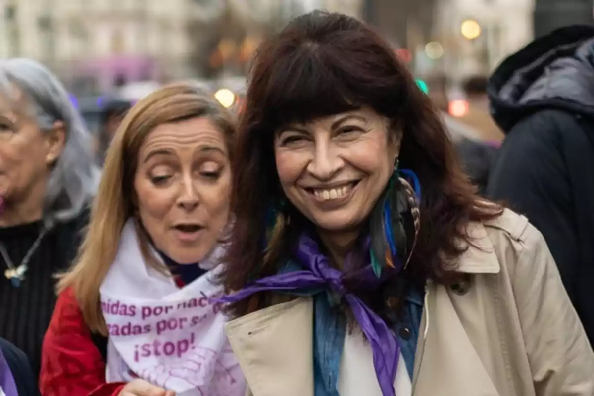 Un grupo de personas sonríe y conversa durante un evento al aire libre, con una mujer en primer plano usando un pañuelo morado y pendientes coloridos.