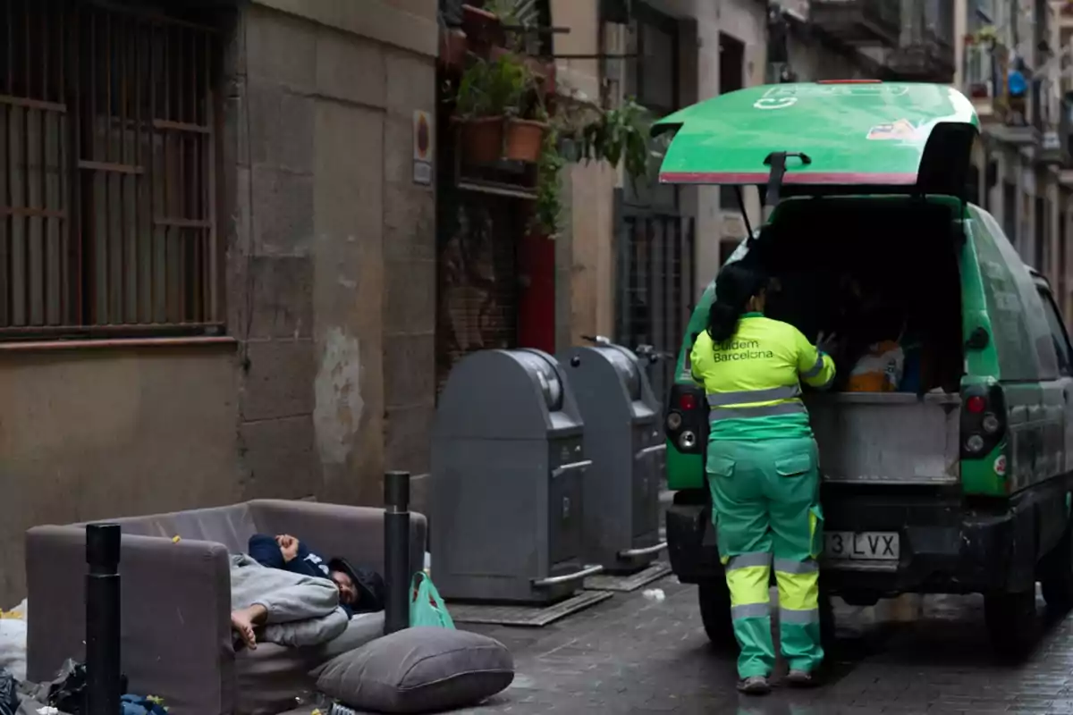 Una persona duerme en un sofá en la calle mientras una trabajadora de limpieza recoge basura de un camión en un entorno urbano. Una persona duerme en un sofá en la calle mientras una trabajadora de limpieza recoge basura de un camión en un entorno urbano.