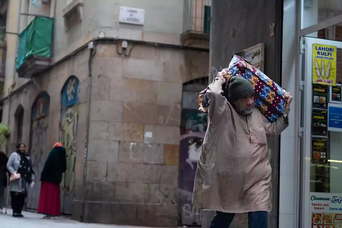 Un hombre mayor con gorro y túnica camina por una calle urbana cargando un paquete grande envuelto en una manta de colores mientras dos mujeres conversan al fondo junto a una pared con grafitis
