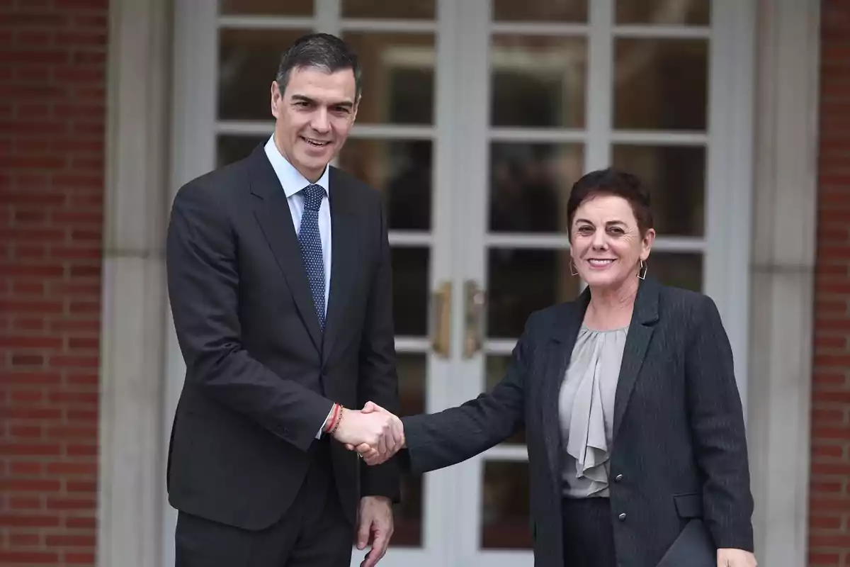 Dos personas vestidas de traje oscuro se dan la mano frente a una puerta de vidrio con marco blanco. Dos personas vestidas de traje oscuro se dan la mano frente a una puerta de vidrio con marco blanco.