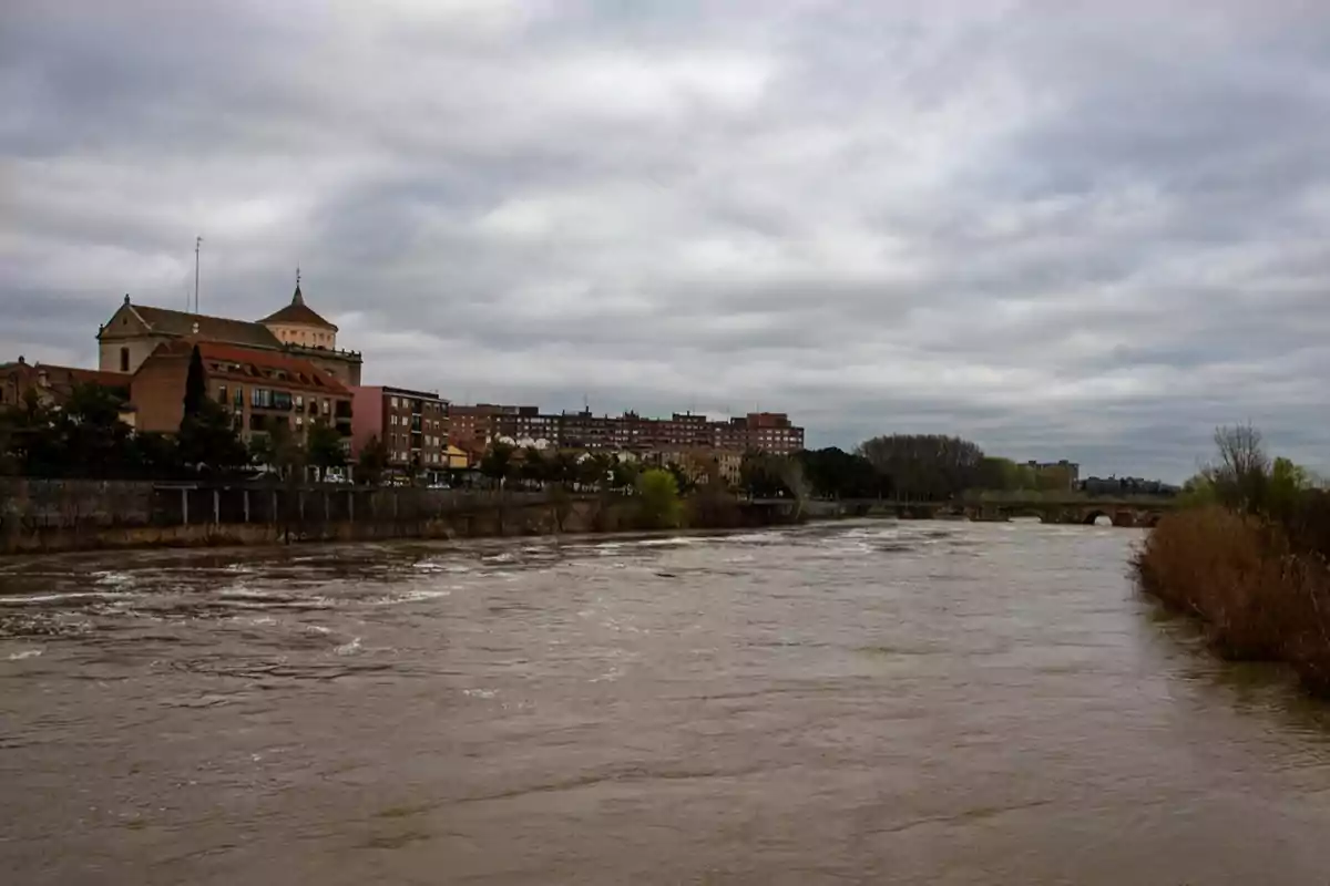 Vista de un río caudaloso con edificios y una iglesia al fondo bajo un cielo nublado.