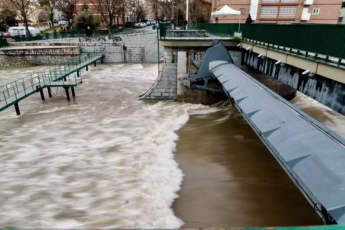 Una estructura de control de agua con compuertas y un canal donde el agua fluye rápidamente, rodeada de barandillas verdes y edificios al fondo.