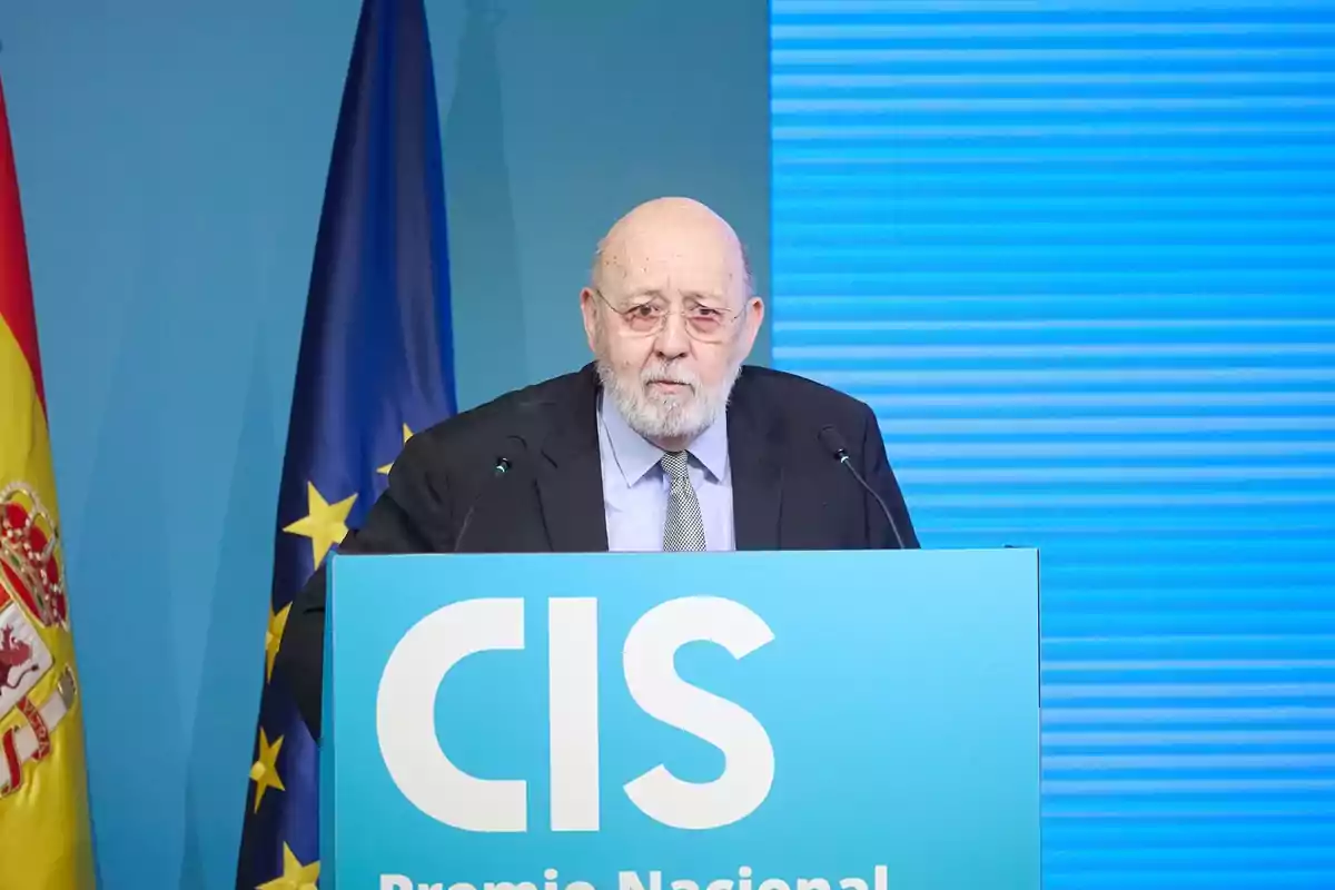 Hombre mayor con barba y gafas hablando en un atril azul con las letras CIS y banderas de España y la Unión Europea al fondo Hombre mayor con barba y gafas hablando en un atril azul con las letras CIS y banderas de España y la Unión Europea al fondo