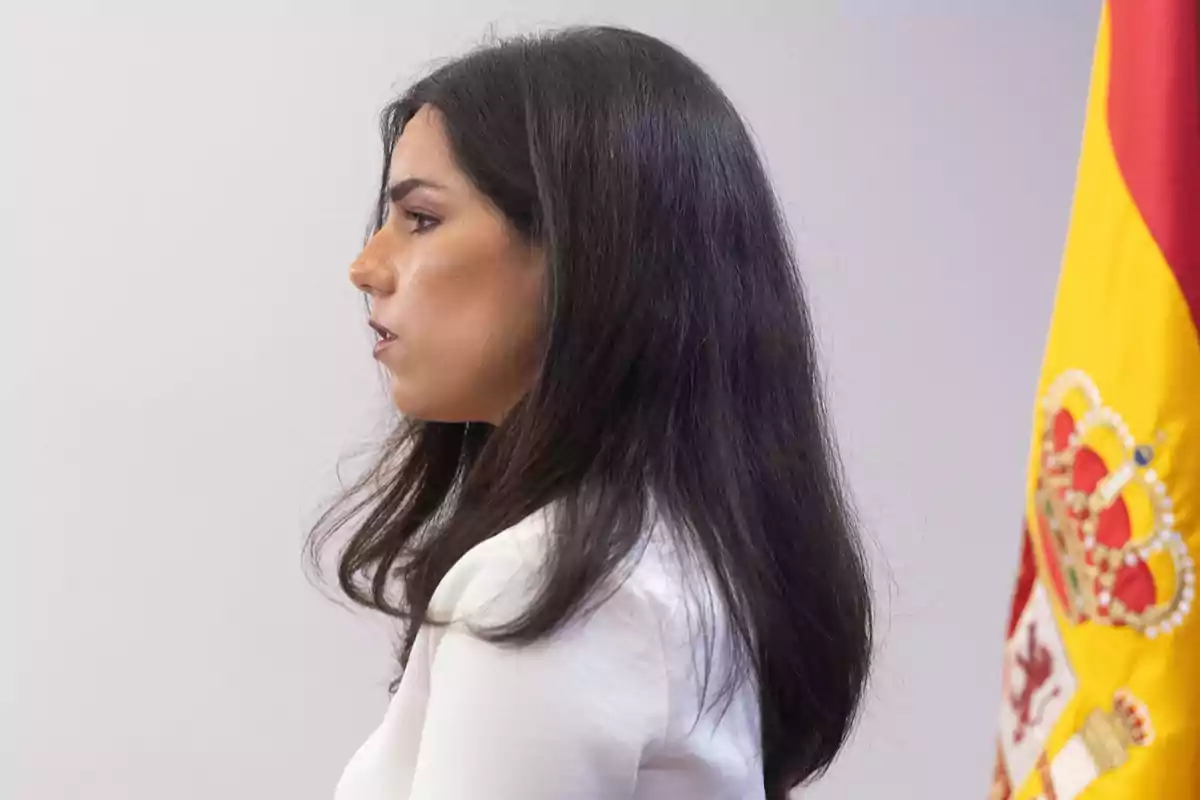 Mujer de perfil con cabello oscuro y largo vestida de blanco junto a una bandera de España. Mujer de perfil con cabello oscuro y largo vestida de blanco junto a una bandera de España.
