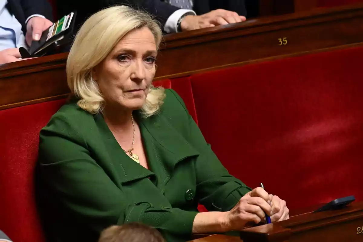 Una persona con cabello rubio y chaqueta verde está sentada en un asiento rojo en un entorno formal. Una persona con cabello rubio y chaqueta verde está sentada en un asiento rojo en un entorno formal.