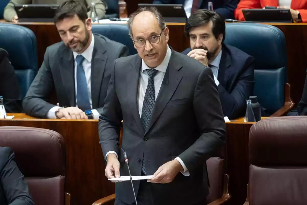 Un hombre de traje y corbata habla en un podio en un entorno formal con otras personas sentadas detrás de él.