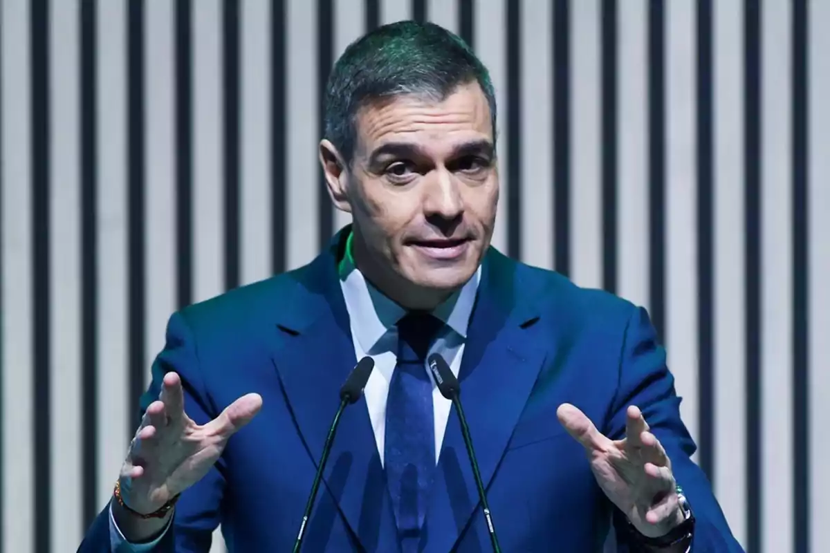 Un hombre en traje azul hablando frente a dos micrófonos con un fondo de líneas verticales. Un hombre en traje azul hablando frente a dos micrófonos con un fondo de líneas verticales.