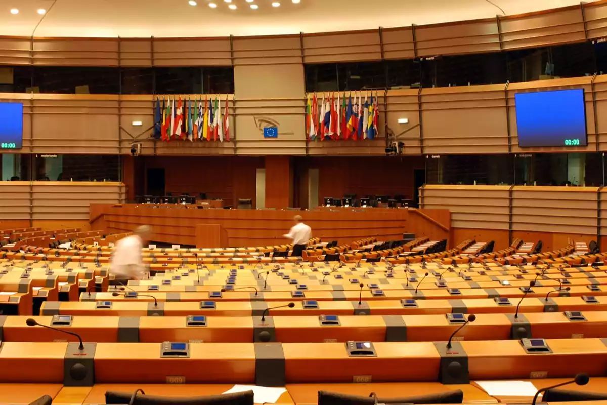 Sala de conferencias moderna con escritorios equipados con micrófonos y pantallas, dos personas caminando y varias banderas de diferentes países al fondo Sala de conferencias moderna con escritorios equipados con micrófonos y pantallas, dos personas caminando y varias banderas de diferentes países al fondo