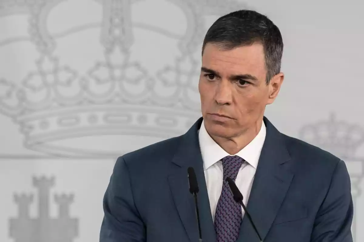 Un hombre en traje azul oscuro y corbata está hablando frente a un micrófono con un fondo que tiene un diseño de corona.