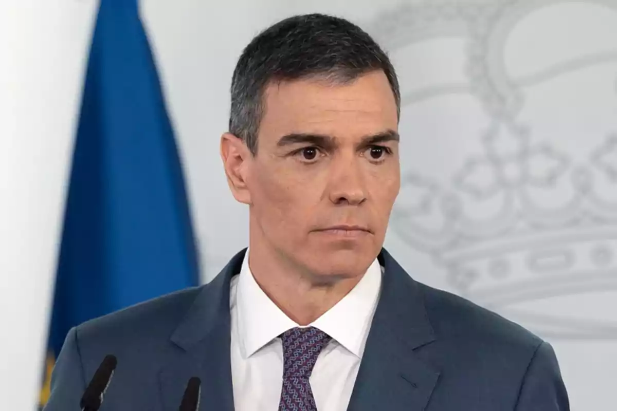 Un hombre con traje azul y corbata, de pie frente a un micrófono con una bandera azul y un fondo con un diseño de corona. Un hombre con traje azul y corbata, de pie frente a un micrófono con una bandera azul y un fondo con un diseño de corona.