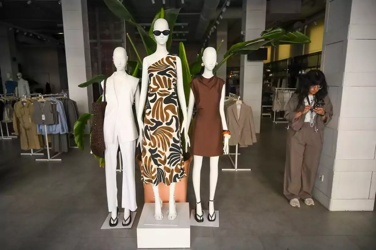 Tres maniquíes con ropa de moda están en exhibición en una tienda de ropa moderna, mientras una persona revisa su teléfono cerca de ellos. Tres maniquíes con ropa de moda están en exhibición en una tienda de ropa moderna, mientras una persona revisa su teléfono cerca de ellos.