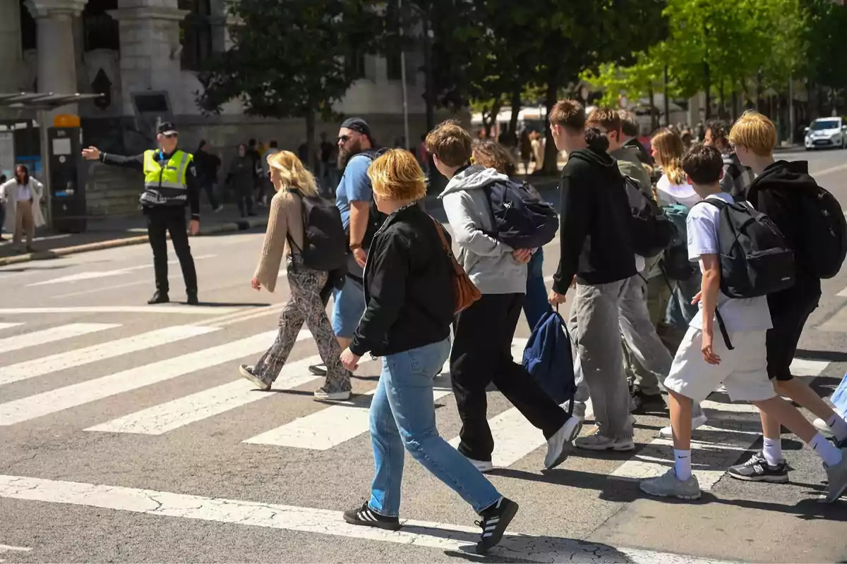 Un grupo de personas cruza un paso de peatones mientras un agente de policía dirige el tráfico en una calle urbana. Un grupo de personas cruza un paso de peatones mientras un agente de policía dirige el tráfico en una calle urbana.