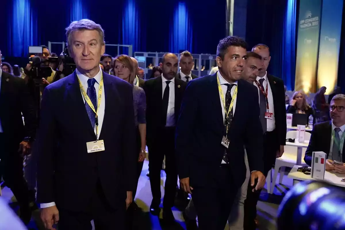 Un grupo de personas vestidas formalmente camina en un evento con iluminación azul y mesas al fondo. Un grupo de personas vestidas formalmente camina en un evento con iluminación azul y mesas al fondo.