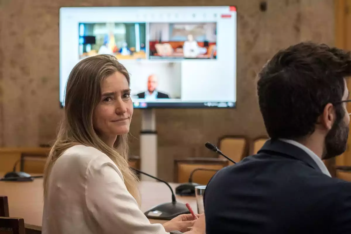 Una mujer sentada en una sala de conferencias mira hacia la cámara mientras un hombre a su lado observa una pantalla grande que muestra una videollamada. Una mujer sentada en una sala de conferencias mira hacia la cámara mientras un hombre a su lado observa una pantalla grande que muestra una videollamada.