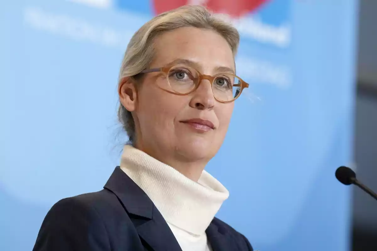 Una persona con gafas y cabello rubio recogido, vestida con un suéter blanco de cuello alto y un saco oscuro, frente a un micrófono y un fondo azul. Una persona con gafas y cabello rubio recogido, vestida con un suéter blanco de cuello alto y un saco oscuro, frente a un micrófono y un fondo azul.
