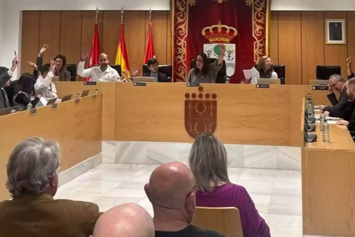 Sesión de un pleno municipal con varias personas levantando la mano para votar, banderas oficiales y un escudo en la pared, mientras algunos asistentes observan desde el público. Sesión de un pleno municipal con varias personas levantando la mano para votar, banderas oficiales y un escudo en la pared, mientras algunos asistentes observan desde el público.