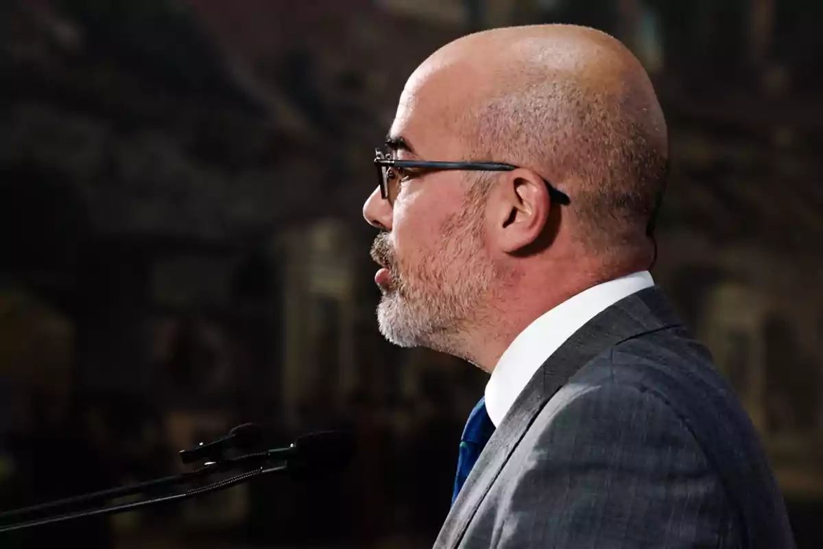 Un hombre calvo con barba y gafas hablando frente a un micrófono. Un hombre calvo con barba y gafas hablando frente a un micrófono.
