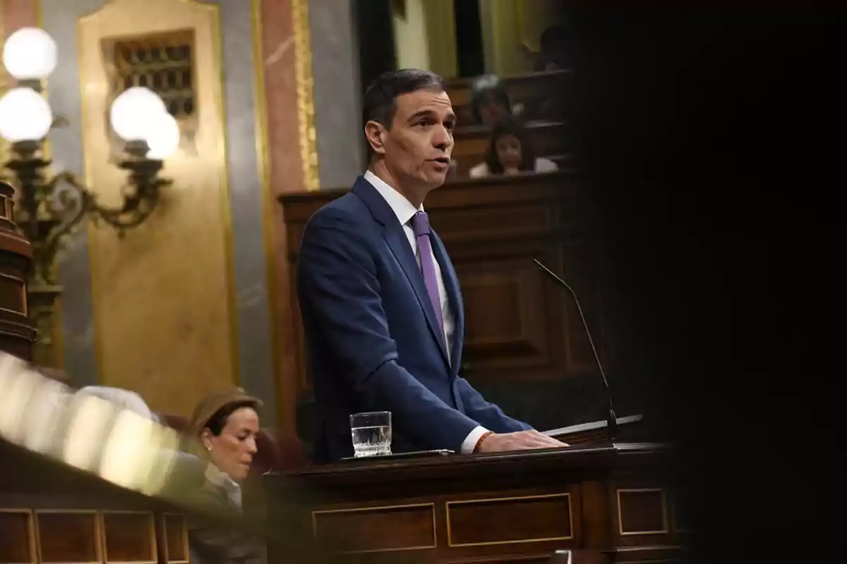 Un hombre con traje azul y corbata morada habla en un atril dentro de un recinto parlamentario con decoración elegante y personas sentadas al fondo Un hombre con traje azul y corbata morada habla en un atril dentro de un recinto parlamentario con decoración elegante y personas sentadas al fondo
